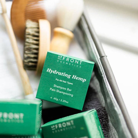 A close-up of a green box of Up Front Cosmetics Hydrating Hemp Shampoo Bar placed inside a metal tray with other hair care items, including a wooden brush and a dark towel.